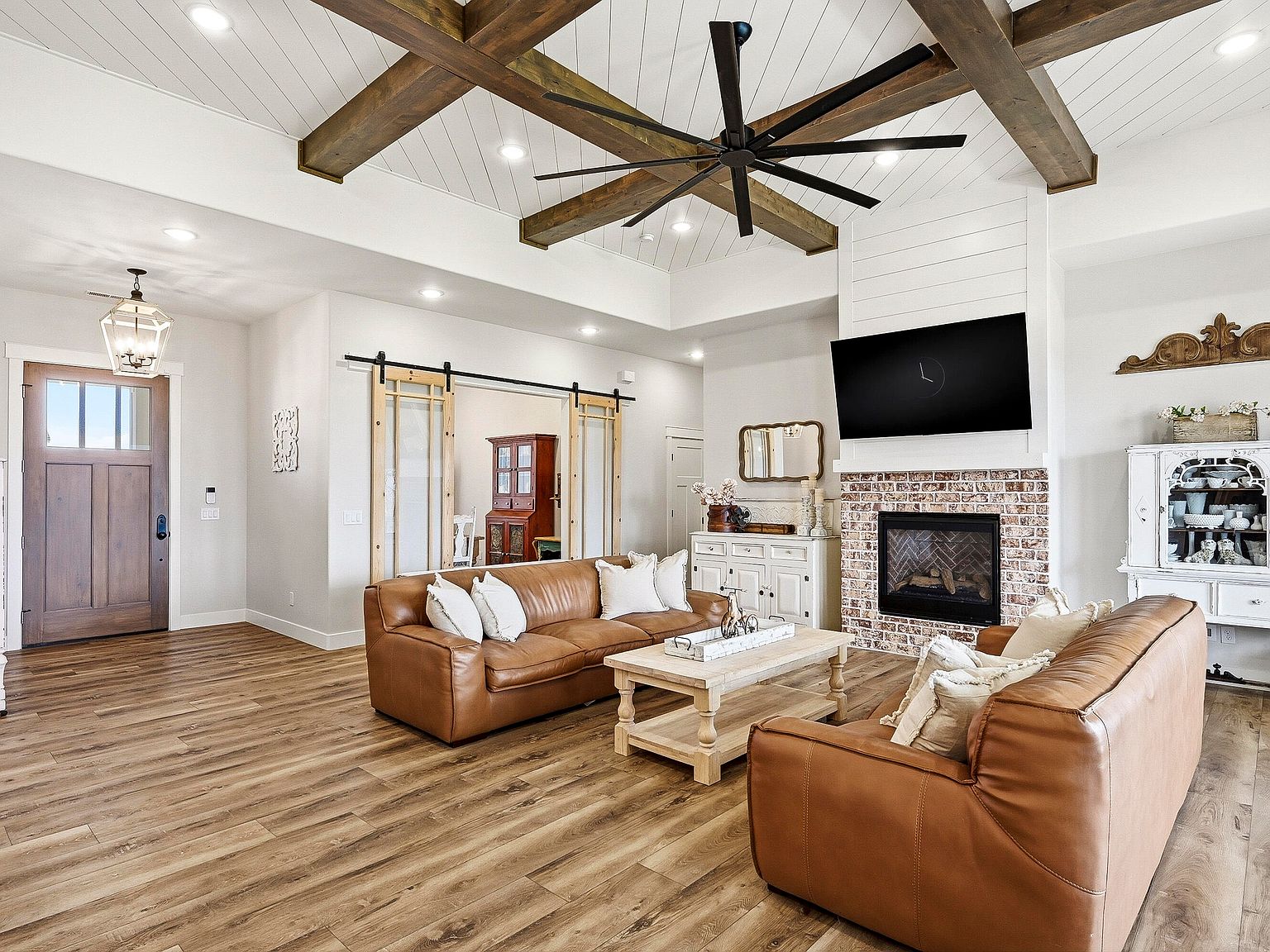 Great room of 7015 W 1300 N with vaulted shiplap ceiling, exposed wood beams, brick fireplace, leather seating, and barn doors leading to the formal dining
